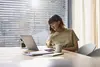 Frau sitzt an einem Tisch, schaut auf einen Laptop und hält eine Tasse. Notizblätter und ein Smartphone liegen auf dem Tisch.