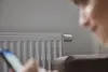 Eine Person schaut auf ein Smartphone, während ein Heizkörper mit einem Thermostat im Hintergrund zu sehen ist.
