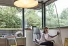 Frau sitzt auf einem modernen Stuhl und berührt ein Fenster, mit einer Stehlampe im Bild.