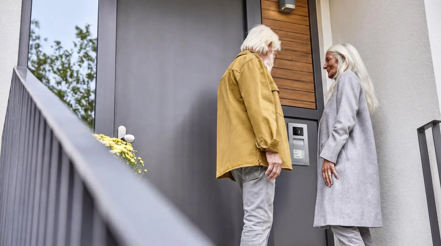 Zwei ältere Personen stehen an einer Eingangstür und betrachten ein Haustelefon oder eine Gegensprechanlage.
