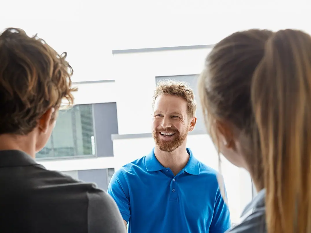 Drei Personen stehen sich gegenüber; ein Mann in einem blauen Poloshirt lächelt freundlich.
