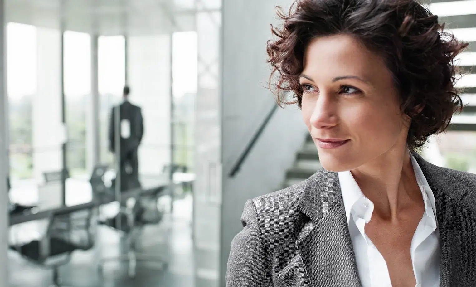 Eine Frau mit lockigem Haar in einem grauen Blazer schaut nachdenklich, während eine Person im Hintergrund steht.