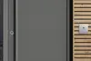 Graue Tür mit einem intercomartigen Klingelsystem und einem kleinen Sichtfenster auf der linken Seite.