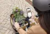 Frau mit Helm steuert ein Lastenrad und nutzt ein Smartphone, um Informationen anzuzeigen. Blumen sind im Radkorb.