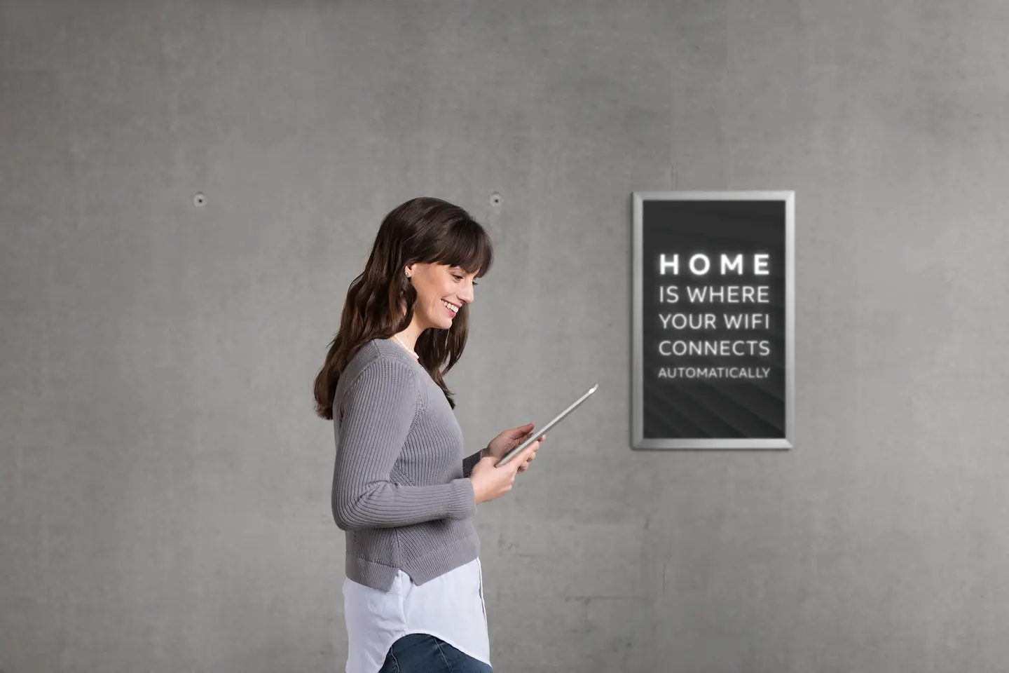 Frau mit Tablet steht neben einem Bild mit dem Text „HOME IS WHERE YOUR WIFI CONNECTS AUTOMATICALLY“.