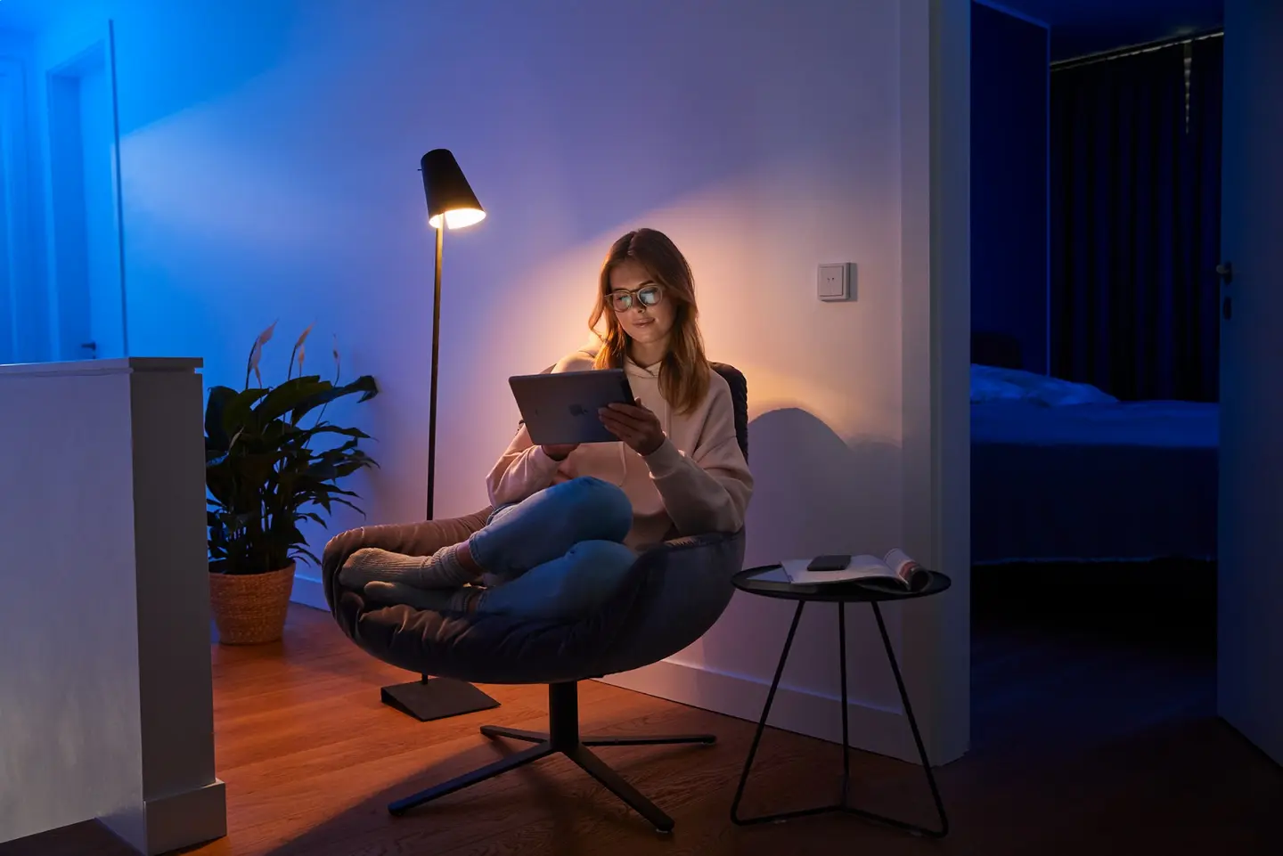 Eine Person mit Brille sitzt entspannt in einem Stuhl und liest auf einem Tablet, beleuchtet von einer Stehlampe.